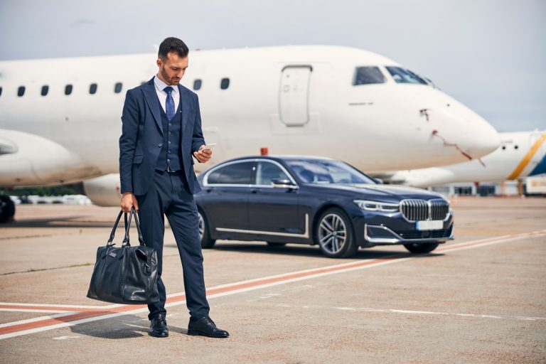 stylish-man-business-suit-looking-his-phone-while-standing-front-car-near-plane
